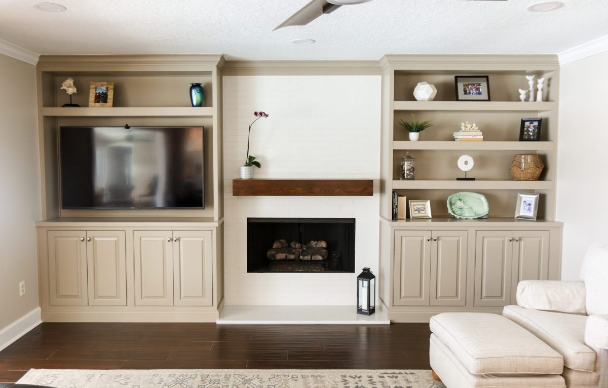 The finished fireplace and media wall: a white brick-pattern tile chimney with a chunky floating wood mantel and firebox at center, flanked by taupe built-in cabinetry &mdash; the television and shelving on one side, open display shelves on the other