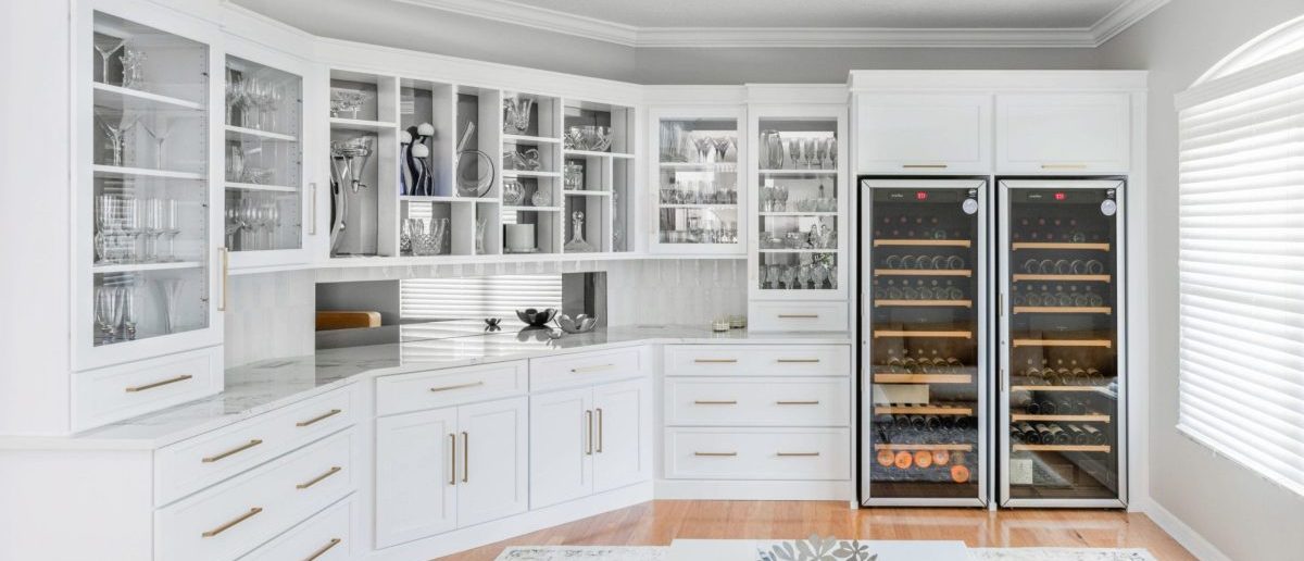 The finished wine bar room: white glass-front display cabinetry filled with crystal stemware wrapping two walls, brass hardware, and two full-height glass-door wine refrigerators built into the cabinetry
