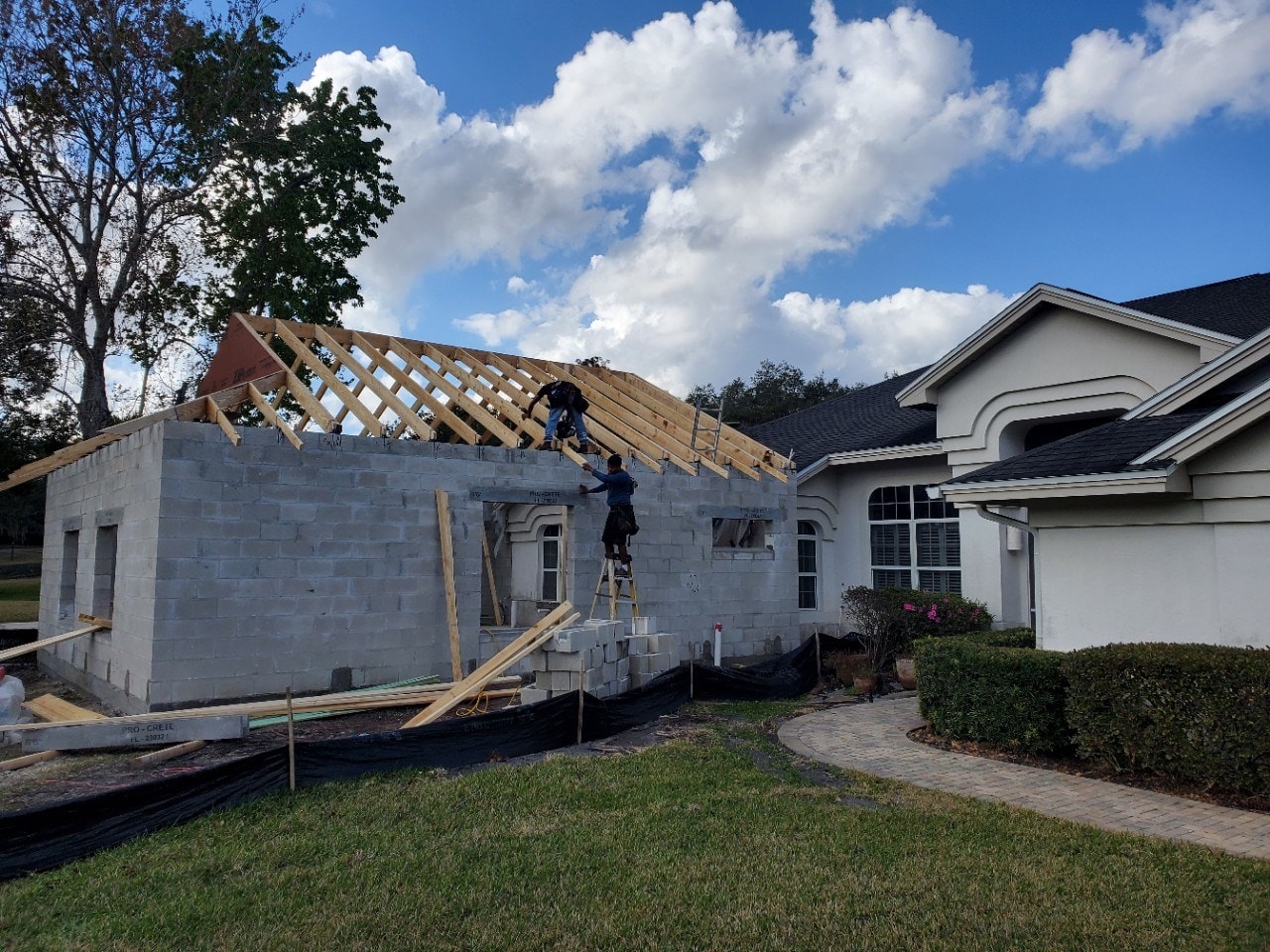 Two workers setting wood roof trusses on the concrete block walls of a new home addition beside a cream stucco home with arched trim and a dark shingle roof, with silt fence and lumber staged in the front yard