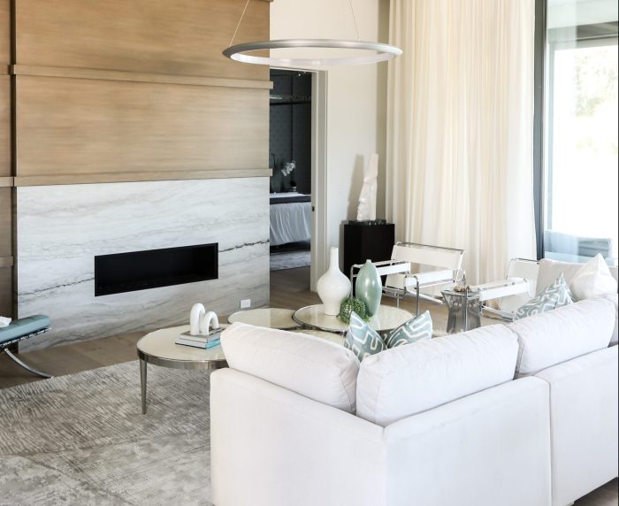 Living room of a whole-residence interior-design project in Orlando &mdash; a linear fireplace set in a travertine-look stone band within a white-oak feature wall, white sectional seating, a ring chandelier and sheer full-height drapes