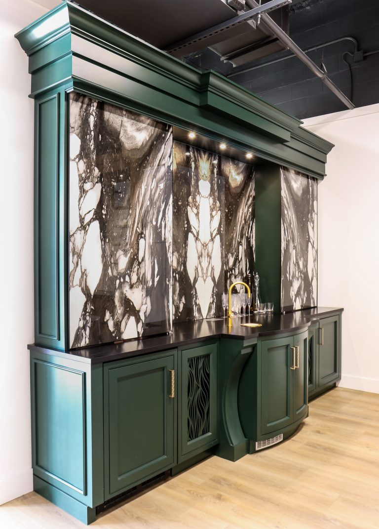 The showroom's bar vignette &mdash; deep-green cabinetry and dramatically veined stone slab panels with a brass faucet, under gallery lighting