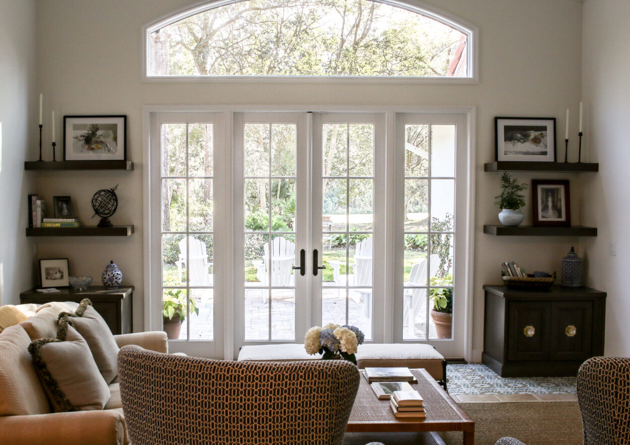 From inside the room: the arched transom and French doors framing the paver terrace, Adirondack chairs, and trees beyond, with floating shelves and console cabinets flanking the opening