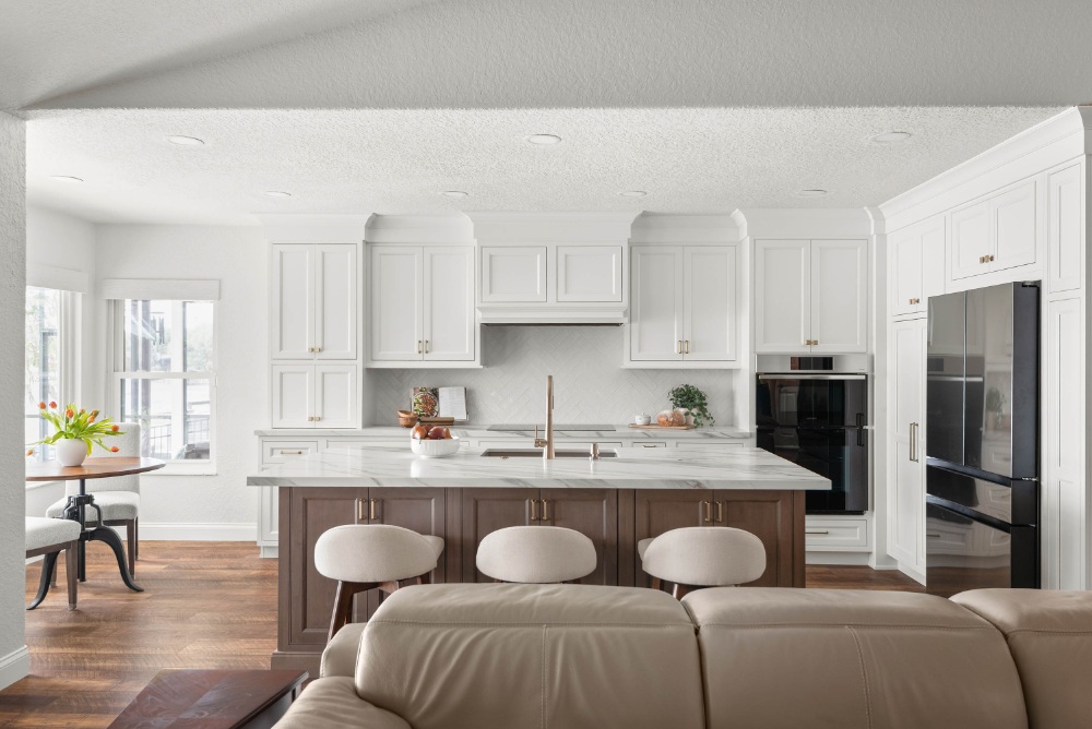 Open-plan remodeled kitchen with a long stained-wood island, three cream counter stools, white cabinetry, herringbone backsplash, and a glass-front refrigerator