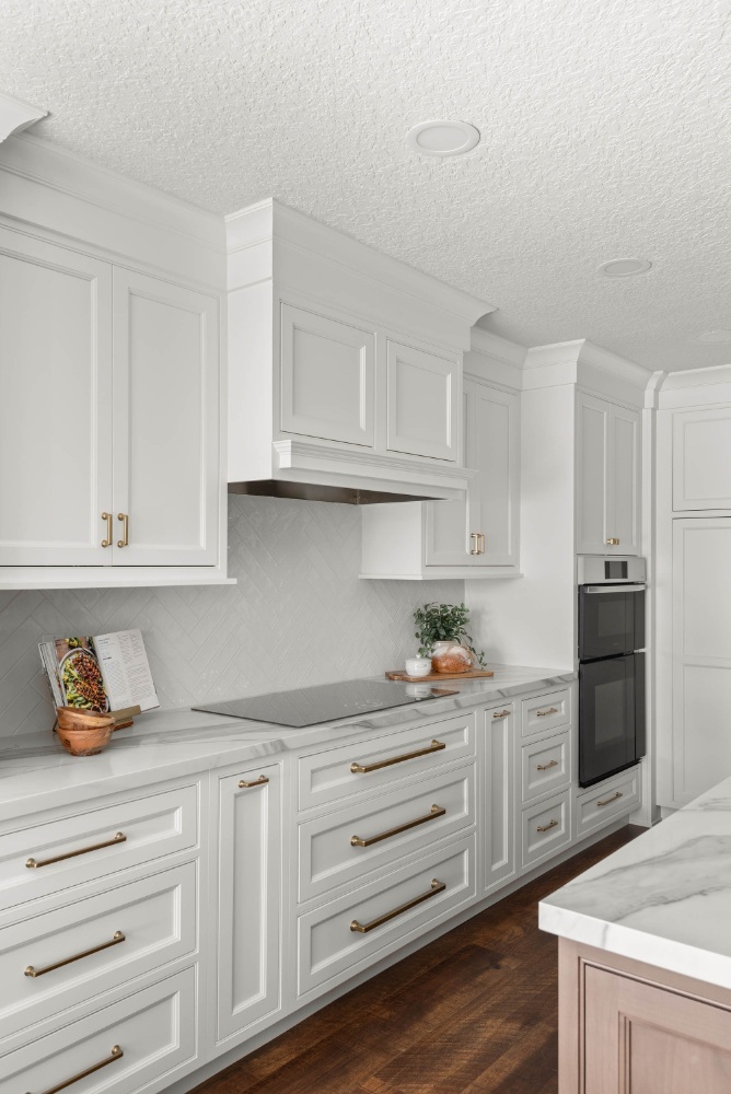 White kitchen cabinetry with brass hardware, a paneled hood, glossy herringbone backsplash tile, and wall ovens