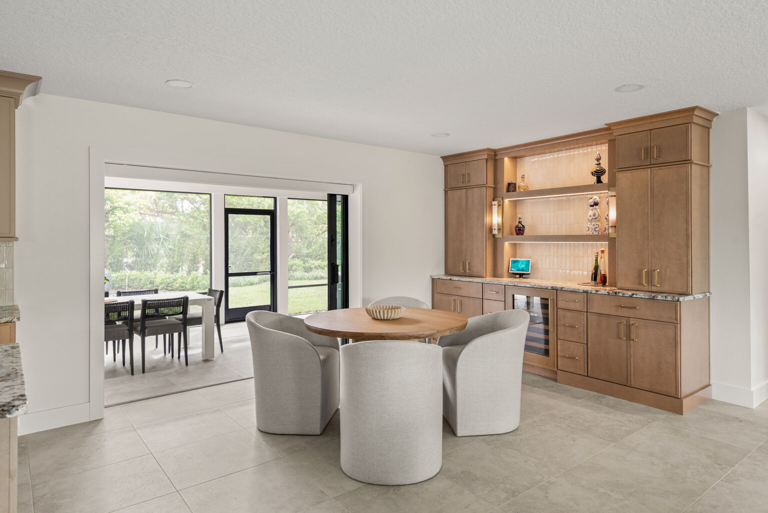 The new eating area and built-in bar after the remodel: a round table with curved chairs, white oak bar cabinetry with lit shelving, and a black-framed glass door opening to the lanai