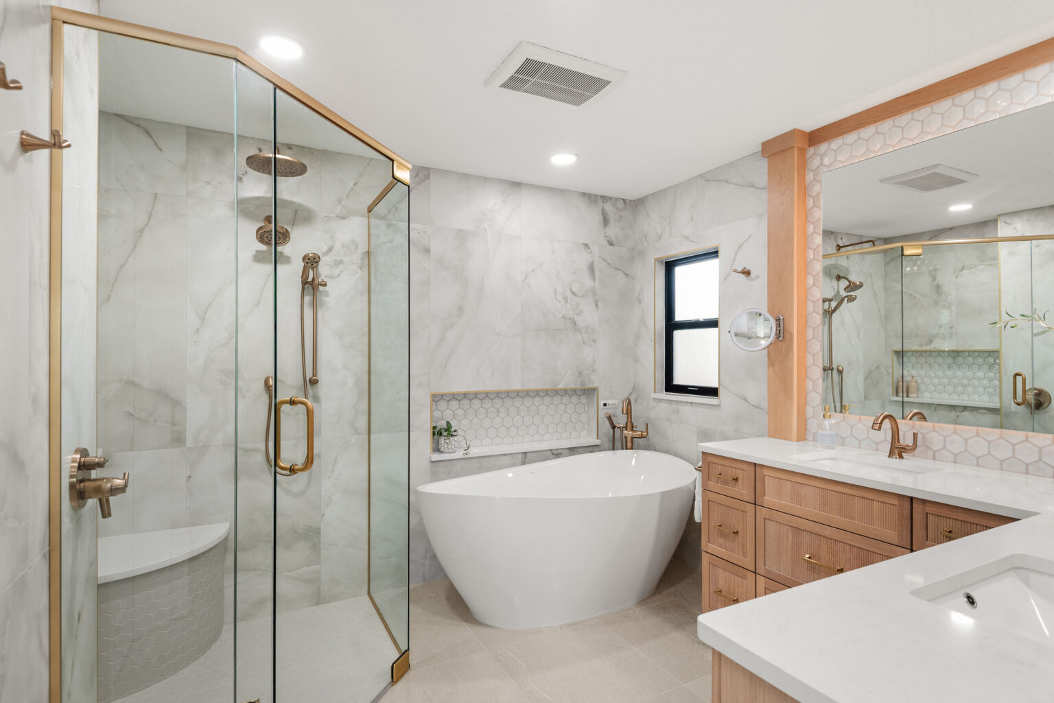 The new primary bathroom created from the spare bedroom: a glass-enclosed shower with brass fittings at left, a freestanding soaking tub beneath a window, and a white oak vanity at right