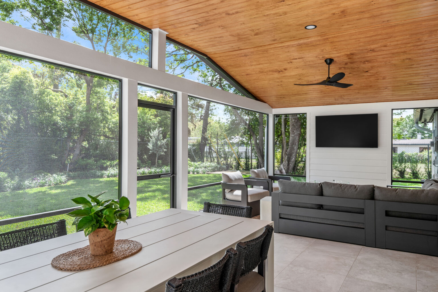 Inside the new screened lanai: a wood-plank ceiling with a fan, a dining table, a sectional sofa and a wall-mounted television looking out to the yard