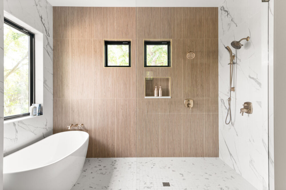 Wet room with a freestanding soaking tub, wood-look tile accent wall, and brass shower fixtures in a Baldwin Park bathroom remodel by KBF Design Gallery