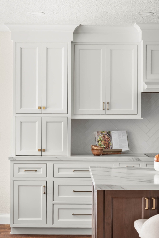 White custom kitchen in a lakefront home remodel by KBF Design Gallery, with floor-to-ceiling shaker storage cabinetry, brass hardware, a herringbone tile backsplash, marble countertops, and a walnut island.