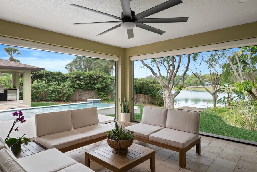 A completed KBF covered outdoor lounge — teak-framed seating under a single wide ceiling fan, looking out past the pool to a separate covered summer kitchen at the left and a lake beyond