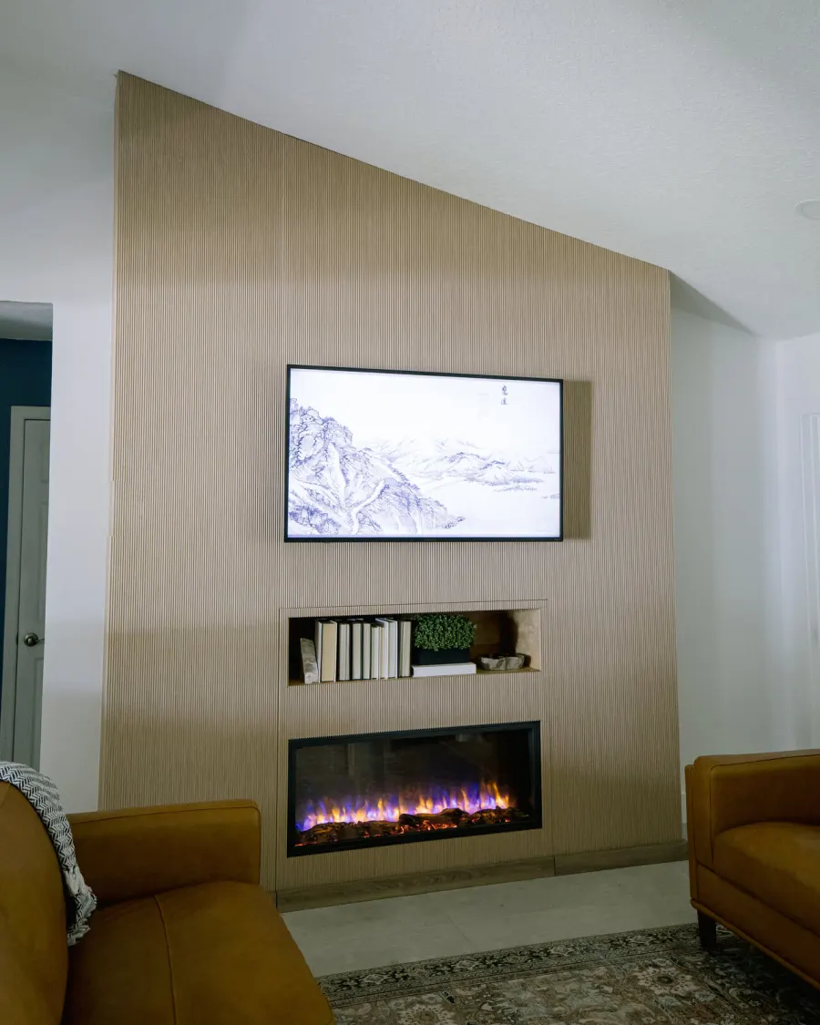 A completed KBF living room — a fluted floor-to-ceiling feature wall carrying a framed wall-mounted display, a lit niche of books and a plant below it, and the top of a linear fireplace at its base