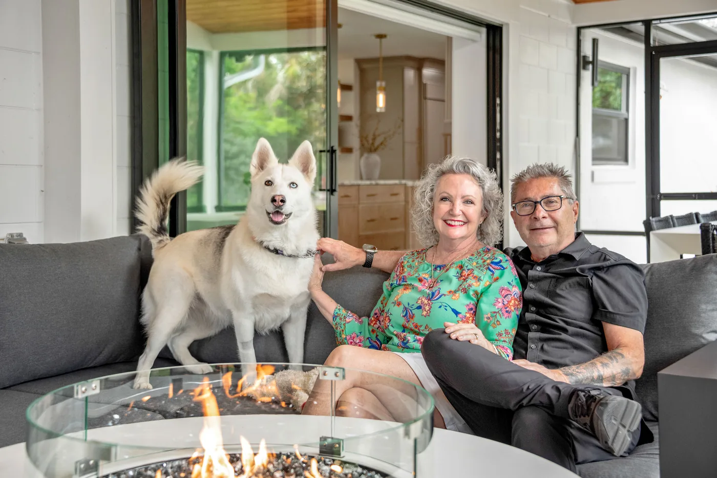 The homeowners with their dog beside the fire table on their new covered patio, the remodeled kitchen beyond