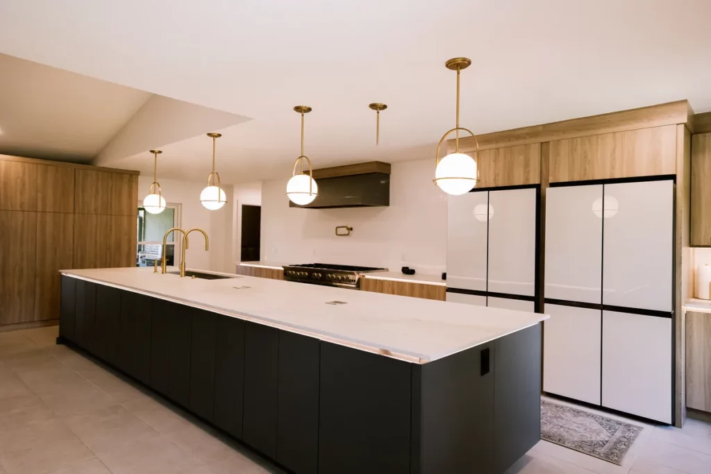 A long charcoal island with a white marble top under brass globe pendants, a professional range beneath a black-and-wood hood, floor-to-ceiling white-oak cabinetry and two panel-front refrigerator columns