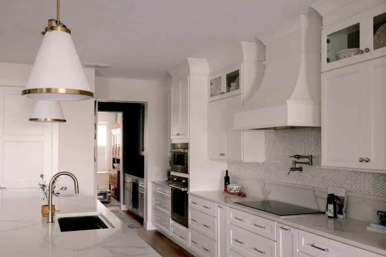 A sculpted plaster hood running to the ceiling above an induction cooktop and wall-mounted pot filler, beside a stacked wall-oven column, with a herringbone marble mosaic backsplash