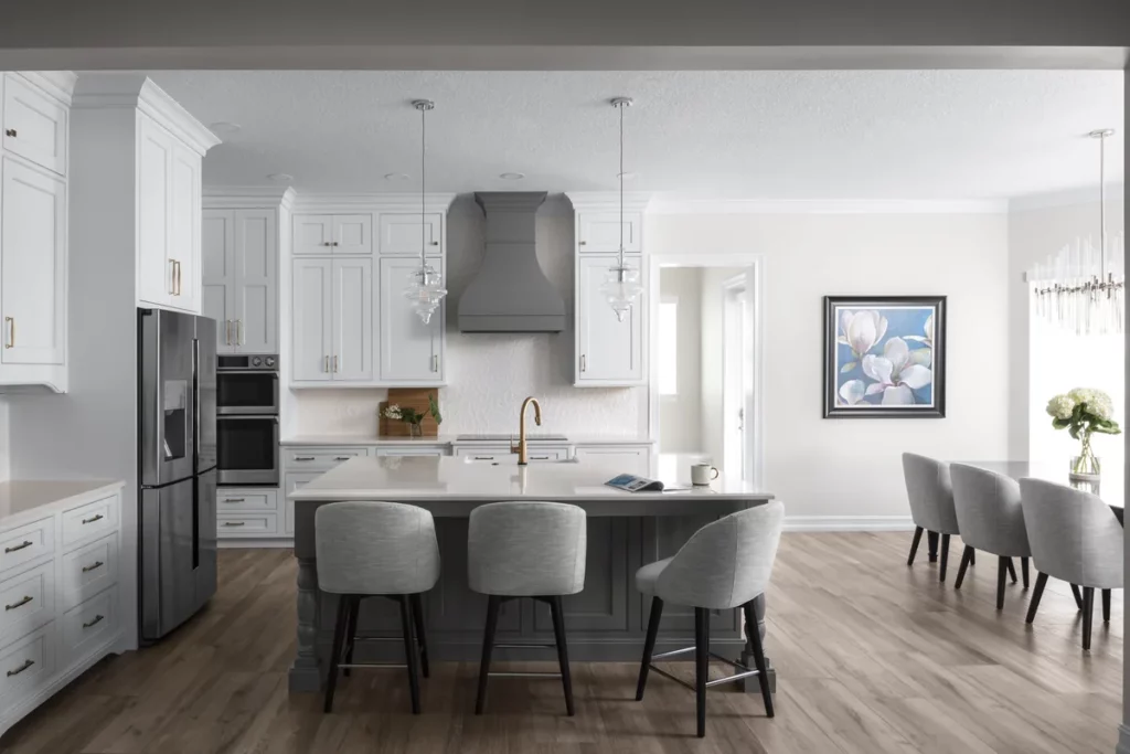 A finished white shaker kitchen with a grey plaster hood, a grey island with three upholstered stools and a brass faucet, opening through to a dining room beyond