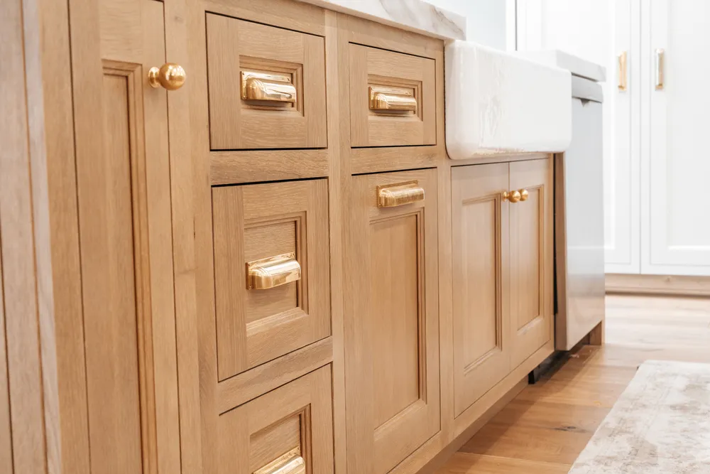 A close-up of white-oak cabinetry with aged-brass bin pulls beside a farmhouse apron sink, in raking light