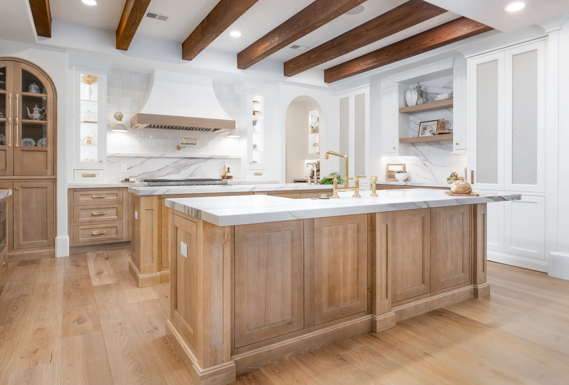 The finished kitchen — a white-oak island with a marble waterfall top, brass fixtures, a plaster hood and exposed wood ceiling beams