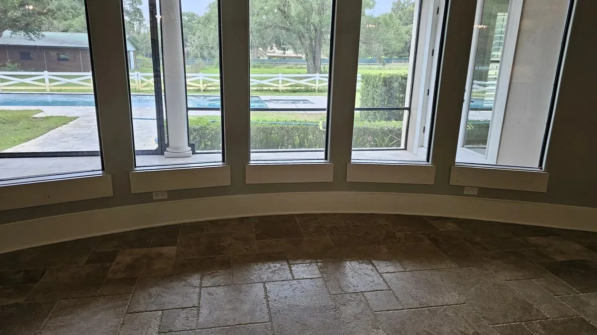 The breakfast room before the remodel: a curved wall of windows with low white sills, travertine floors, and the pool and white cross-rail paddock fence outside