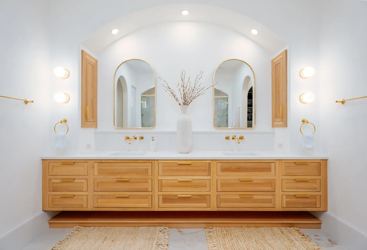 The same Windermere primary bath as chapter 03 &mdash; a floating double white-oak vanity under arched brass mirrors, marble counter and aged-brass wall taps