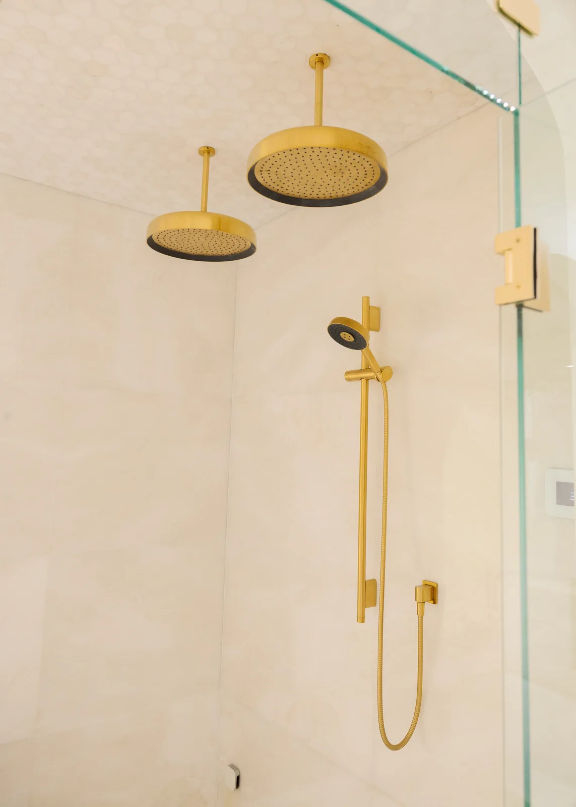 Inside the Winter Springs steam shower by KBF Design Gallery: twin brass rain heads overhead against a honed hex mosaic ceiling, a brass slide-bar hand shower on the polished large-format tile wall, the recessed control panel at the right edge beyond the frameless glass