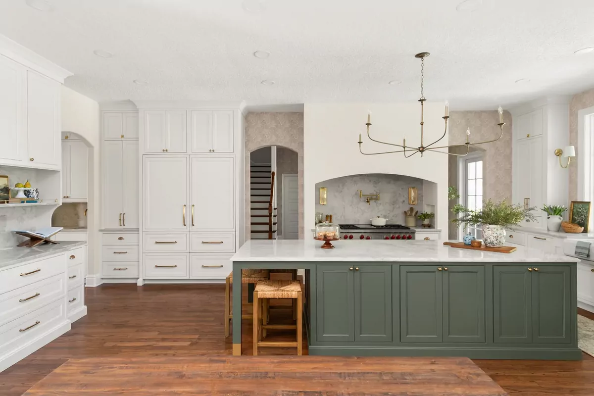 A kitchen after remodeling by KBF Design Gallery: a sage-green island under a thick white marble top with rush-seat stools, a marble-clad arched range alcove with a brass pot filler, floor-to-ceiling white paneled cabinetry, a brass chandelier, a refinished oak floor and the arched opening to the staircase at the centre