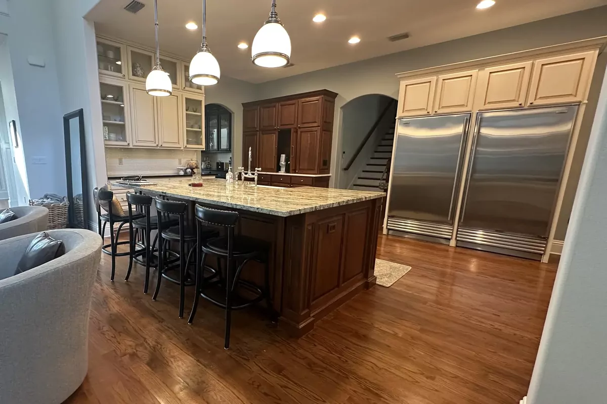 A kitchen photographed before remodeling: a dark cherry island under speckled granite with black bar stools, cream upper cabinets, a stained wood hutch, two stainless refrigerators side by side, and an arched opening to the staircase beyond