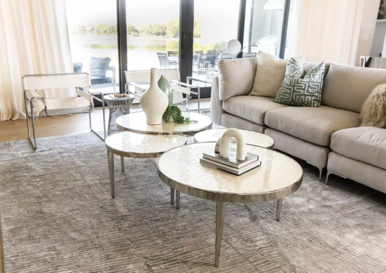 A KBF interior-design living room — nesting stone-top tables, a linen sectional and Wassily chairs held against a full wall of glass onto the water