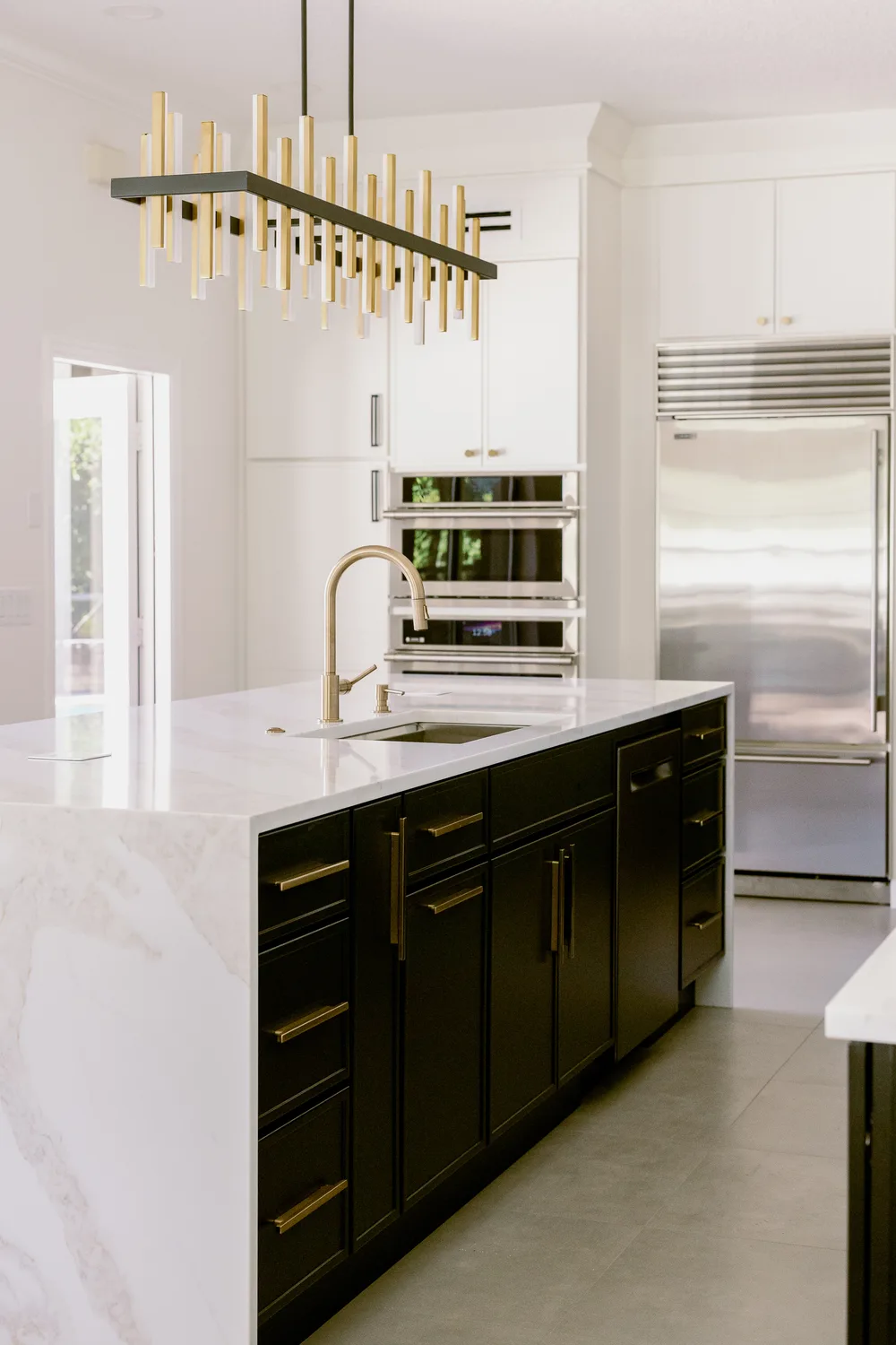 Dommerich Estates kitchen remodel in Maitland — a black center island with a white waterfall-edge counter, a brass-and-black linear chandelier, white cabinetry, wall ovens and a stainless refrigerator