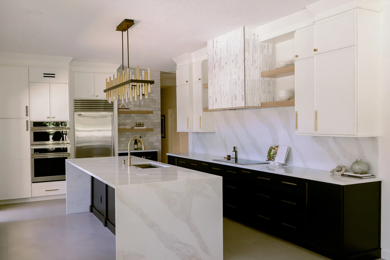 Dommerich Estates kitchen remodel in Maitland &mdash; a marble waterfall island under the brass-and-black linear chandelier, black perimeter cabinets with brass pulls, wall ovens and a stainless refrigerator