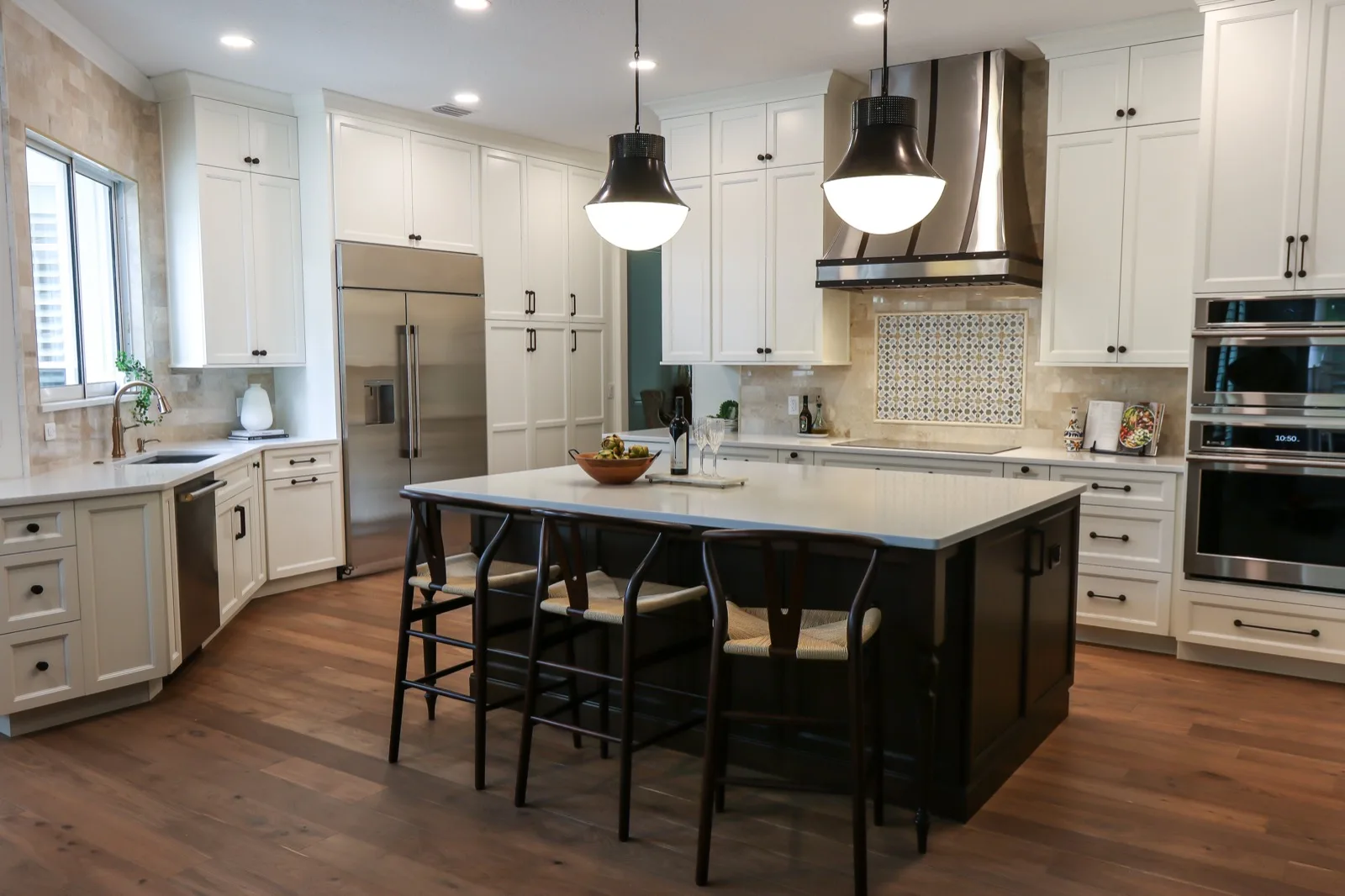 Maitland kitchen remodel, the full room — a dark-wood island with wishbone counter stools under two black dome pendants, white perimeter cabinetry, a strapped stainless chimney hood over a blue-patterned tile inset, a stainless refrigerator and double wall ovens, and travertine-toned backsplash tile