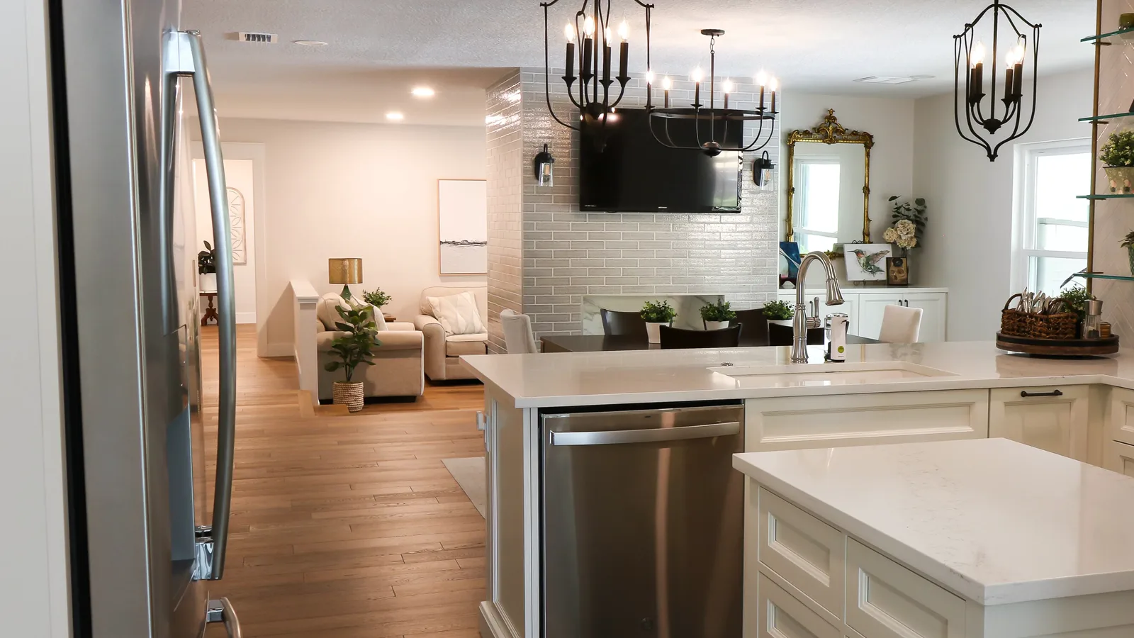The completed kitchen of a Maitland whole-home renovation — a white quartz peninsula, a stainless chimney hood, brass-and-glass shelving on herringbone tile, and wood floors running through to the living room