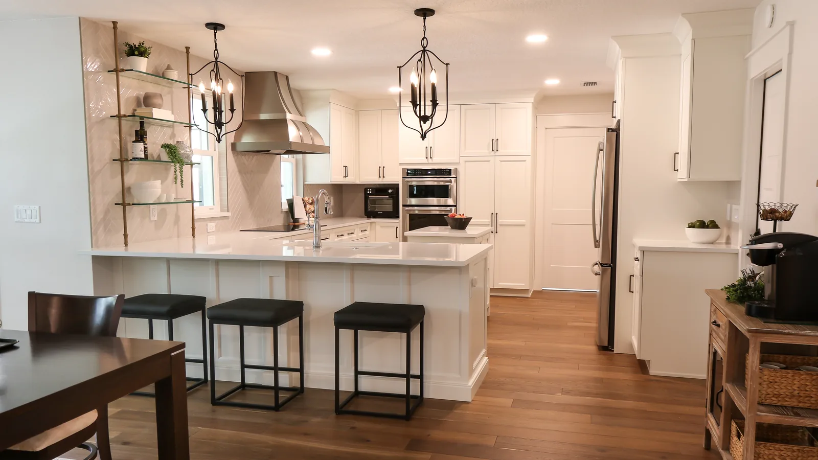 The same Maitland kitchen after the whole-home renovation — a white quartz peninsula with three black counter stools open to the dining room, a stainless chimney hood over the cooktop, brass-and-glass shelving on herringbone tile, and wood floors running through to the living room