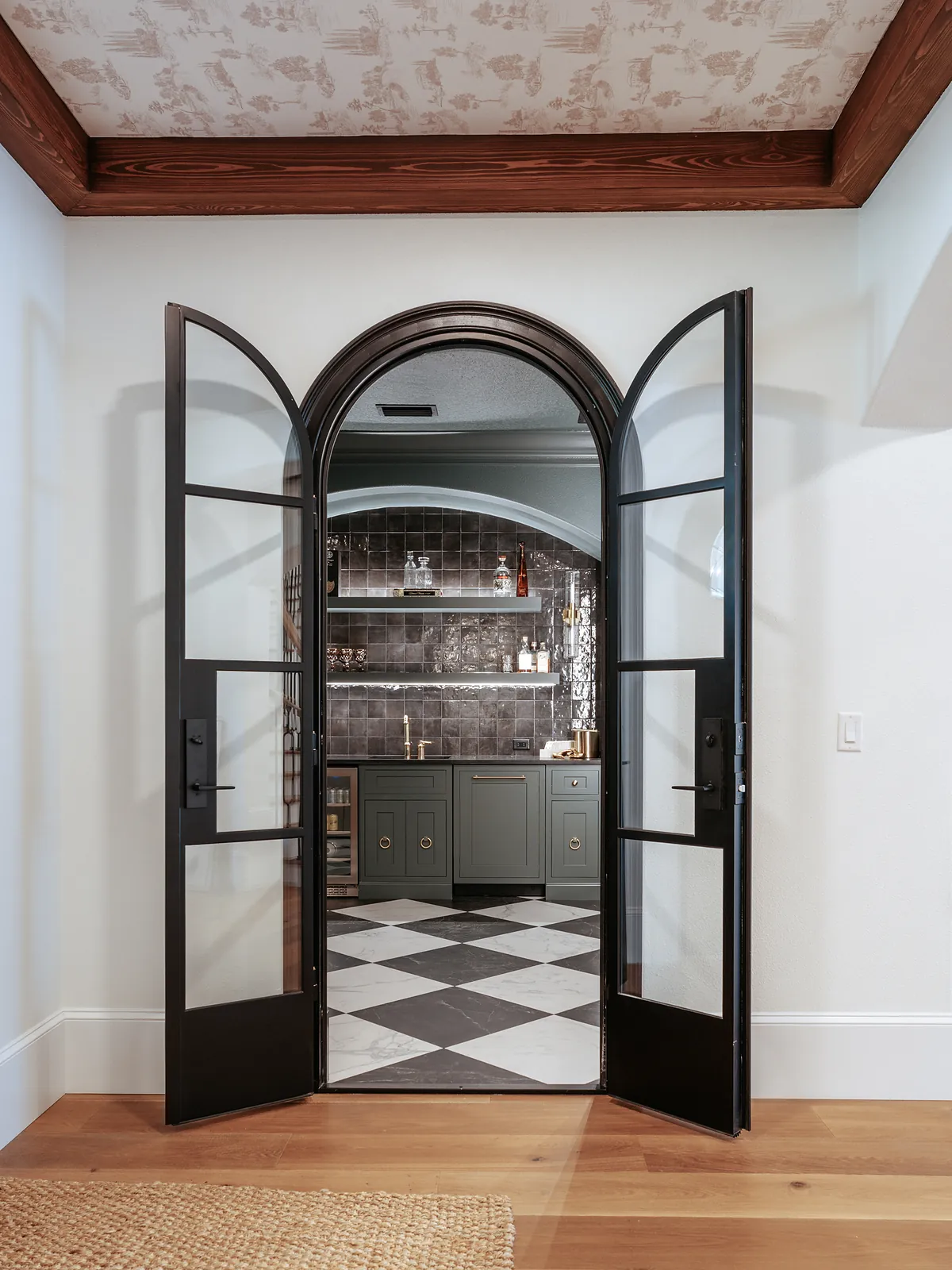 The finished wine bar behind open arched steel-and-glass doors — green cabinetry with brass ring pulls, a black glazed tile backsplash, floating shelves, a brass faucet and a black-and-white marble checkerboard floor