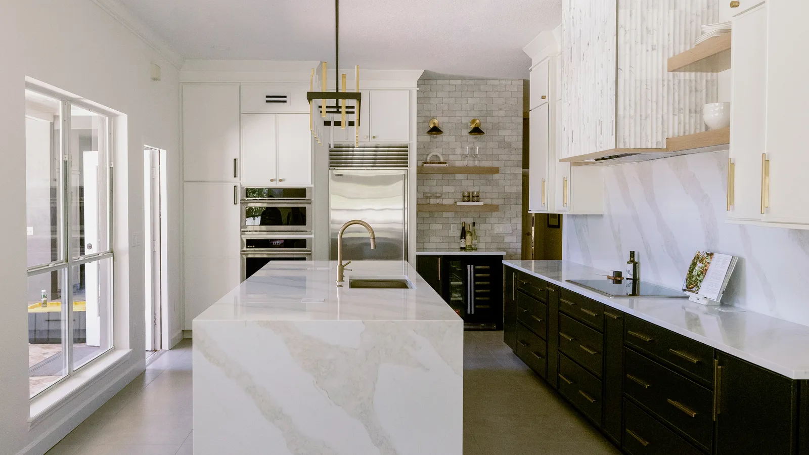 The same Maitland kitchen after remodeling: white cabinetry with brass hardware, a marble slab hood surround and backsplash, dark lower cabinets along the right-hand run, a stone-topped island with a brass faucet, and a lit tile niche beside the refrigerator