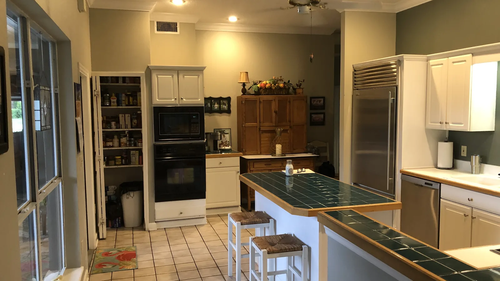 The Maitland kitchen before remodeling: cream raised-panel cabinets, dark green tile counters and a tiled peninsula bar with two stools, black double wall ovens, an open pantry and terracotta floor tile