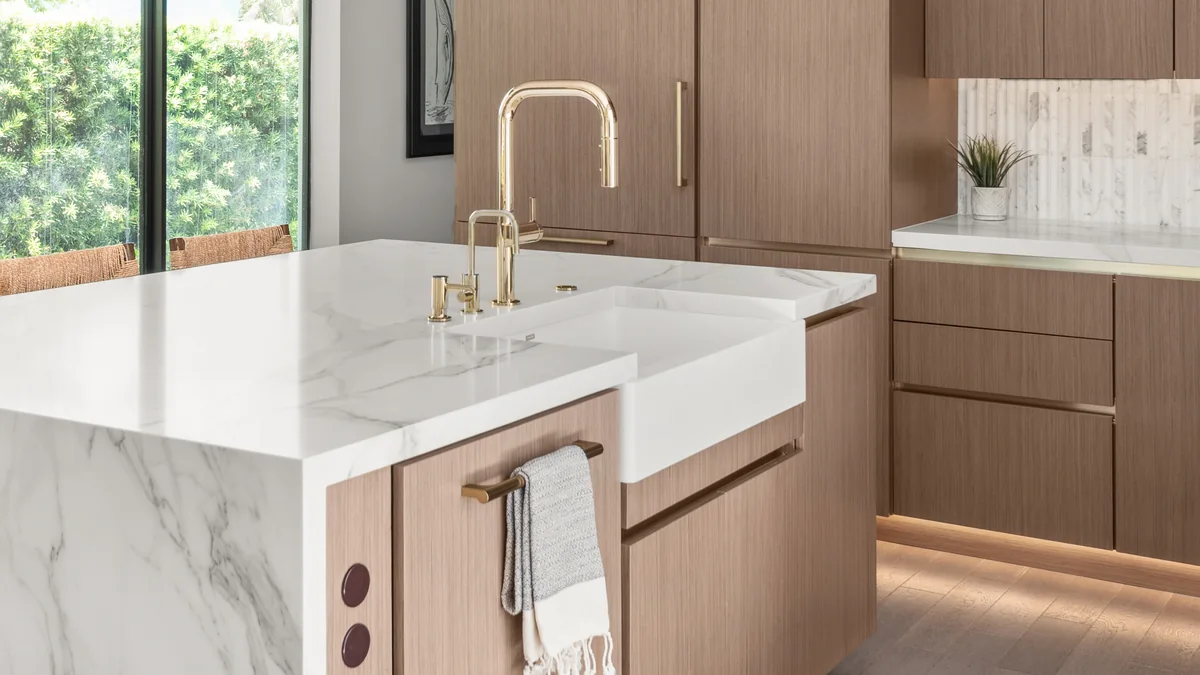 A Studio KBF kitchen at completion: a marble-veined quartz waterfall island with an apron-front sink, a brass faucet, pop-up outlets set into the island side, and wood-grain slab cabinetry beyond