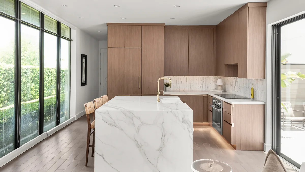 A completed Studio KBF kitchen in Winter Park seen from the dining end: wood-grain slab cabinetry, a marble-veined quartz island, a stacked-mosaic backsplash and the window wall alongside