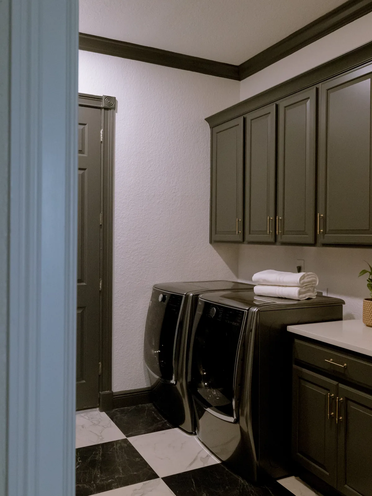 The same laundry room at completion: deep-olive wall cabinets with brass pulls carried up to a crown, a white quartz counter over olive base cabinets, a graphite washer and dryer, and the same checkerboard floor