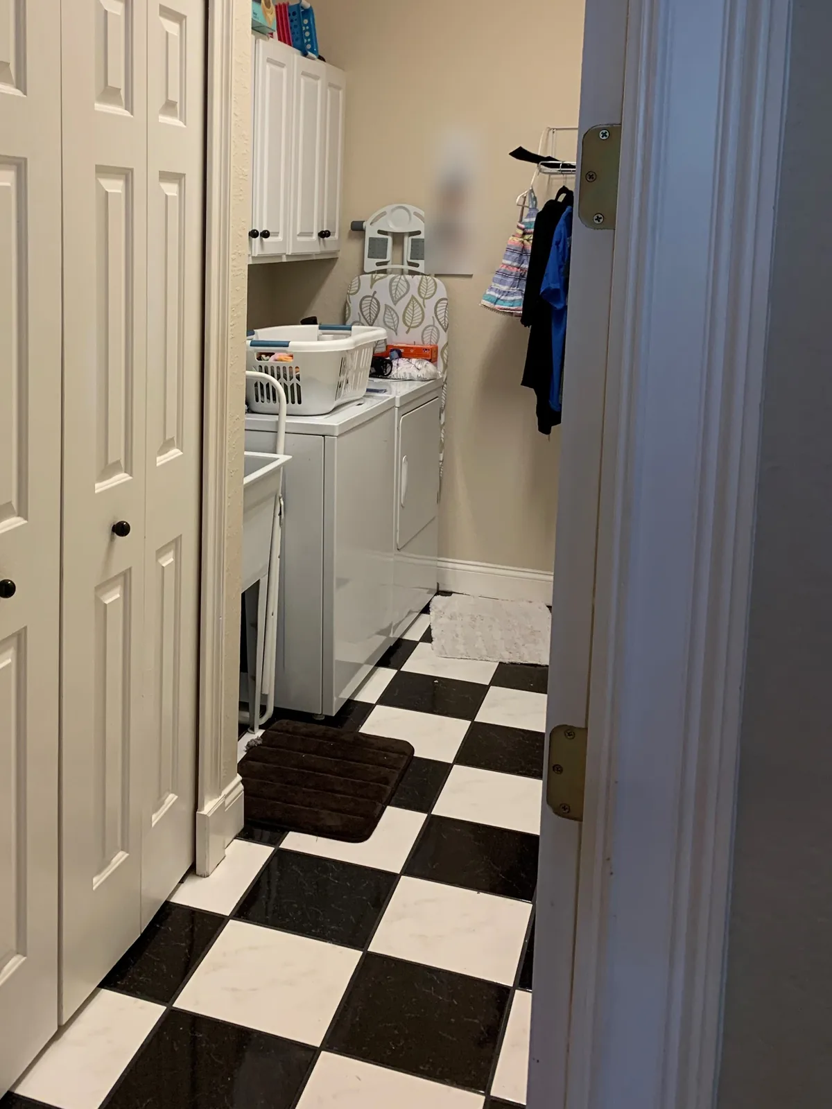 The laundry room before remodeling, seen from the hallway: white builder cabinets over a white washer and dryer, beige walls, a wire shelf, and a black-and-white checkerboard floor