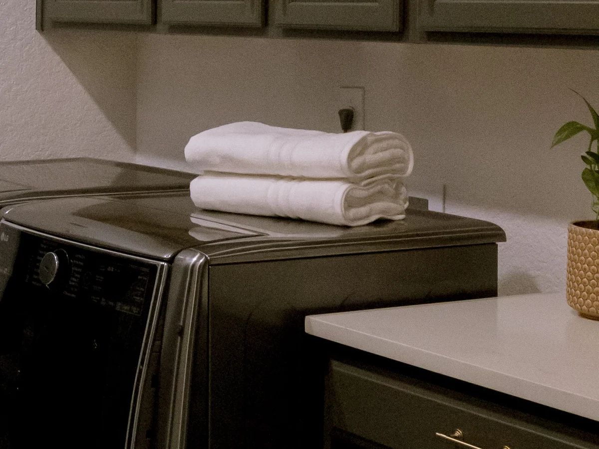 Finish detail from the same laundry: folded towels on the graphite washer, the white quartz counter and olive base cabinets with slim brass bar pulls, under the olive wall cabinets