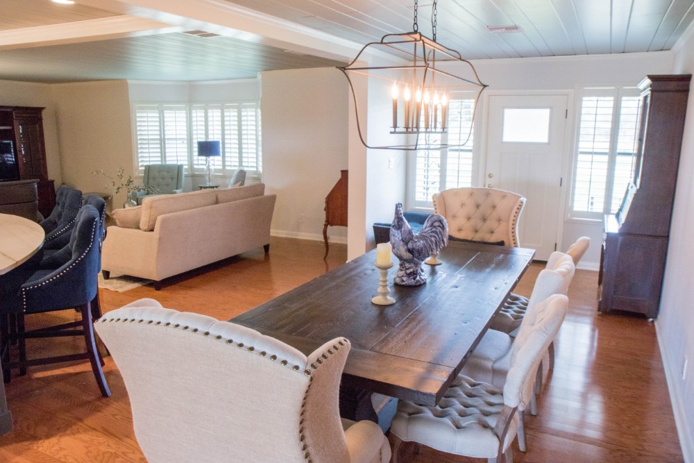 Open dining and living area after remodeling with a farmhouse dining table, upholstered chairs, an iron lantern chandelier, and plantation shutters
