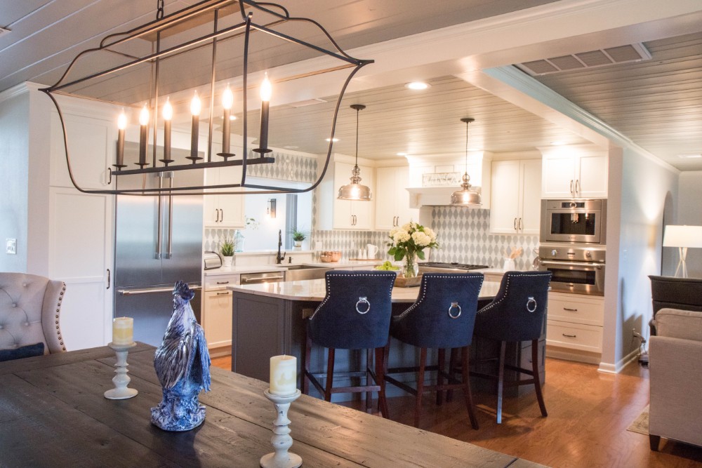 Open-concept modern farmhouse kitchen after remodeling, with white perimeter cabinets, a gray island with navy counter stools, a patterned backsplash, painted plank ceilings, and an iron lantern chandelier over the dining table