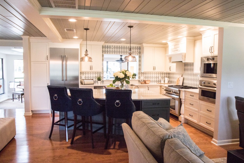 Open-plan view from the living area after remodeling: large gray island with navy counter stools, patterned backsplash, professional range, wall ovens, and a stainless refrigerator under painted plank ceilings