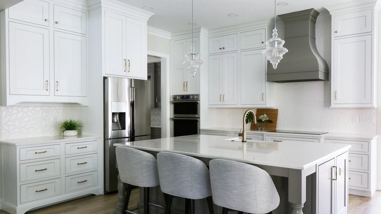 White inset kitchen with a shaped grey range hood, glass pendants over a quartz island with upholstered stools, and a panelled refrigerator &mdash; Celebration kitchen remodel by KBF