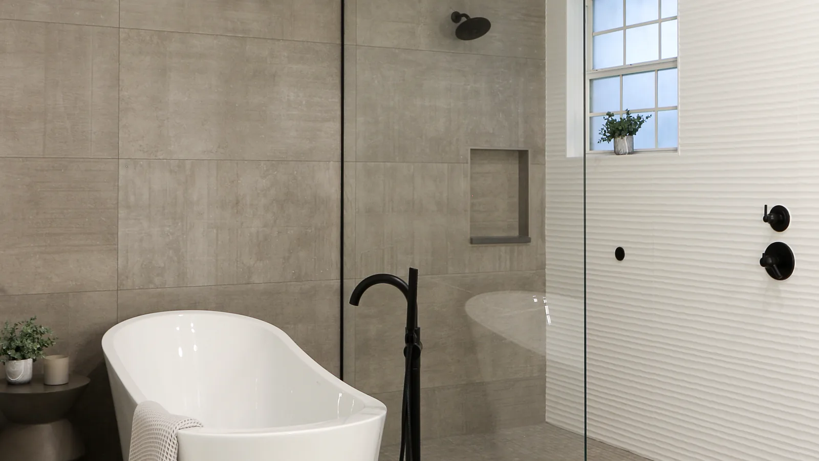 The same Oviedo primary bath after the complete remodel: a freestanding white soaker tub with a matte-black floor-mounted filler beside an open glass-panel shower, the same divided-light window now set over textured white tile