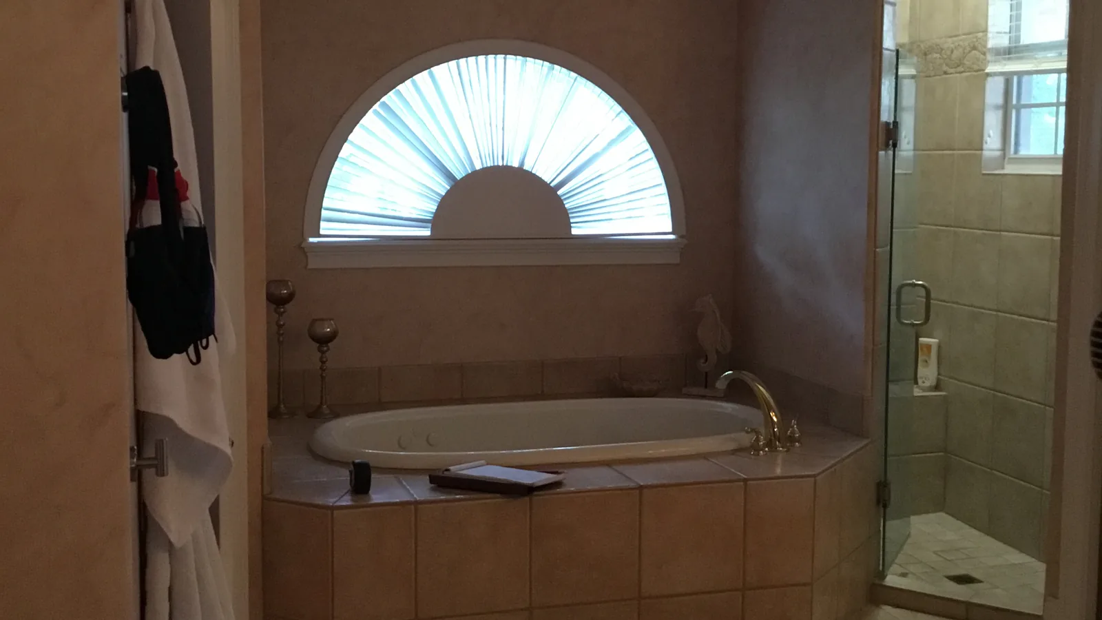 The Oviedo primary bath before remodeling: a drop-in garden tub on a beige tile deck below an arched half-moon window with a fan blind, next to a framed glass shower with a small divided-light window inside