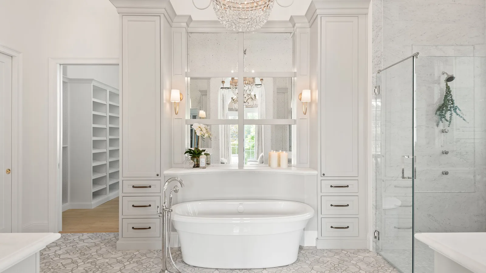 The same Windermere primary bath after the complete remodel: a freestanding oval soaking tub centered between two white cabinetry towers, below an antiqued-mirror paneled surround with sconces and a crystal chandelier