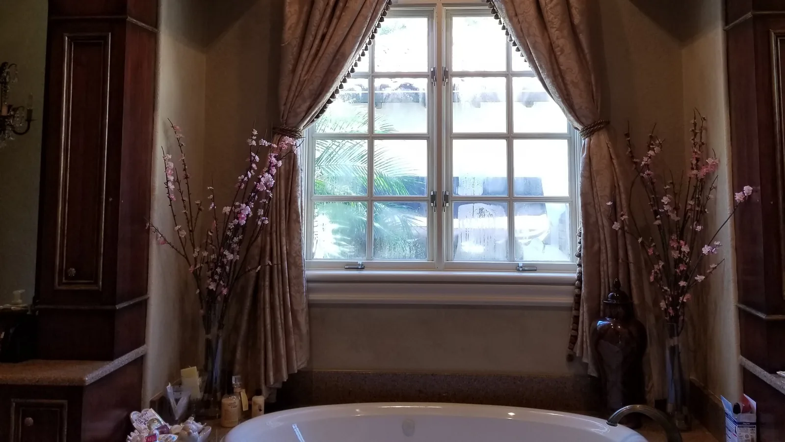 The Windermere primary bath before remodeling: a drop-in jetted tub on a speckled granite deck inside an arched alcove, below a divided-light window with silk draperies, between two dark wood cabinetry towers