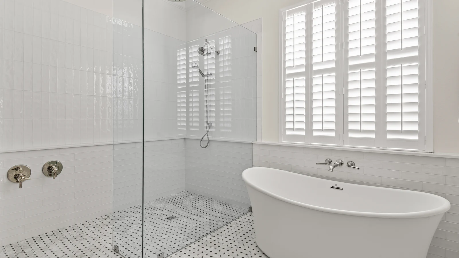The same Winter Park primary bath after remodeling: a white freestanding soaking tub with wall-mounted polished-nickel filler and a frameless glass walk-in shower with basketweave mosaic floor, below the same plantation-shuttered window