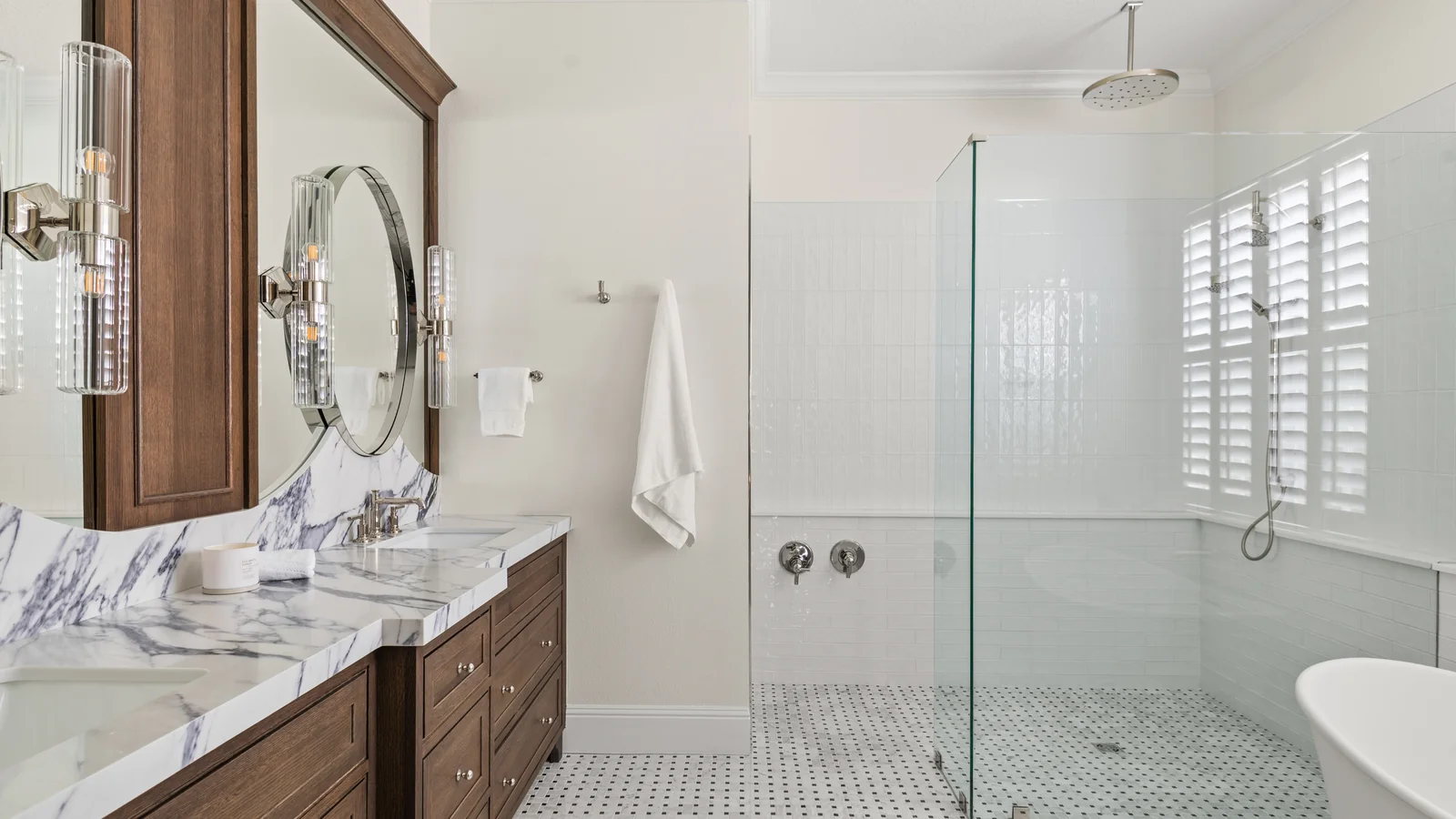 The same Winter Park primary bath after remodeling: a stained-oak double vanity under marble with an oval mirror and fluted-glass sconces, the frameless walk-in shower and freestanding tub beyond
